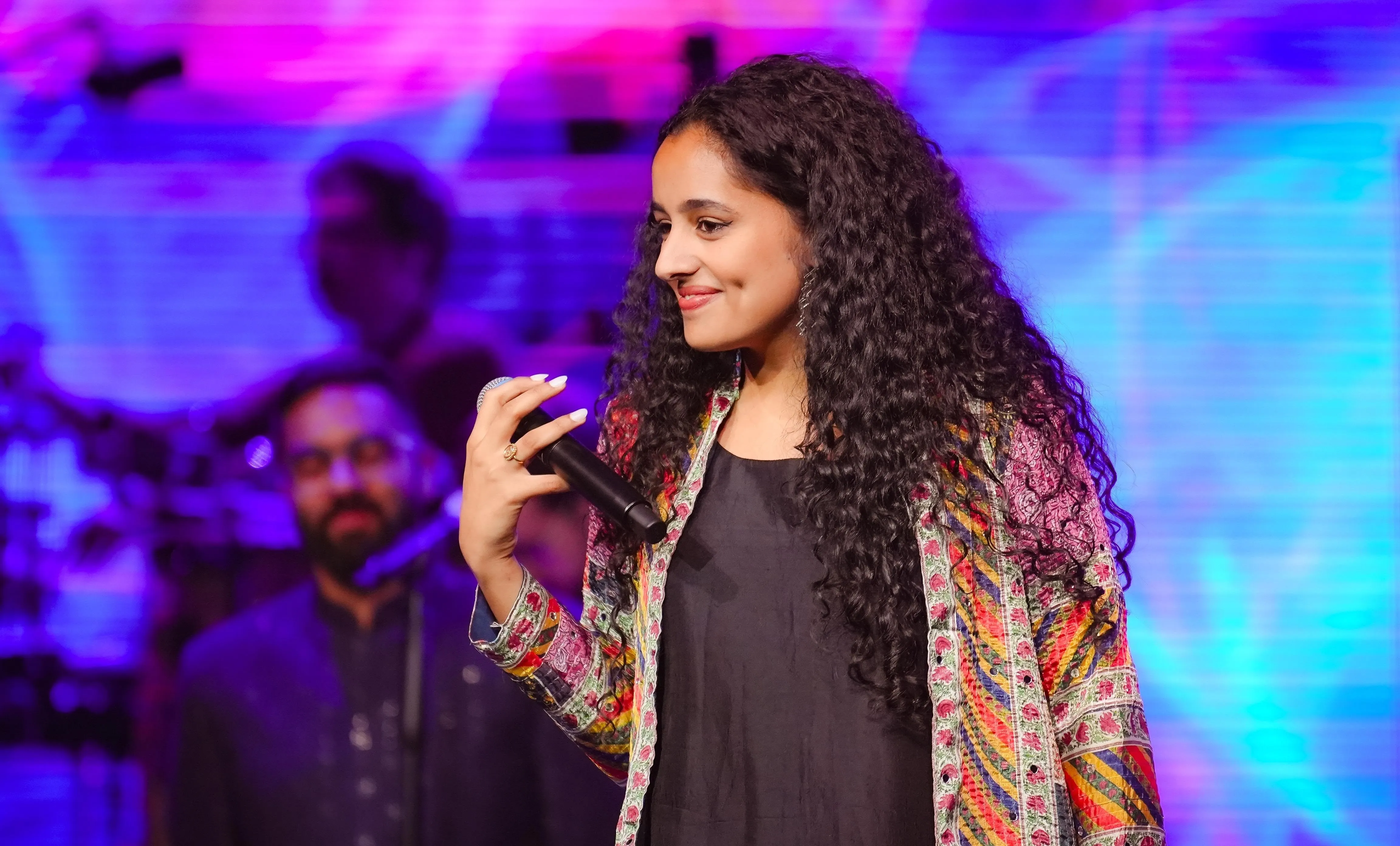 Sindhu Vasudevan on stage with a microphone, mid-performance under stage lights