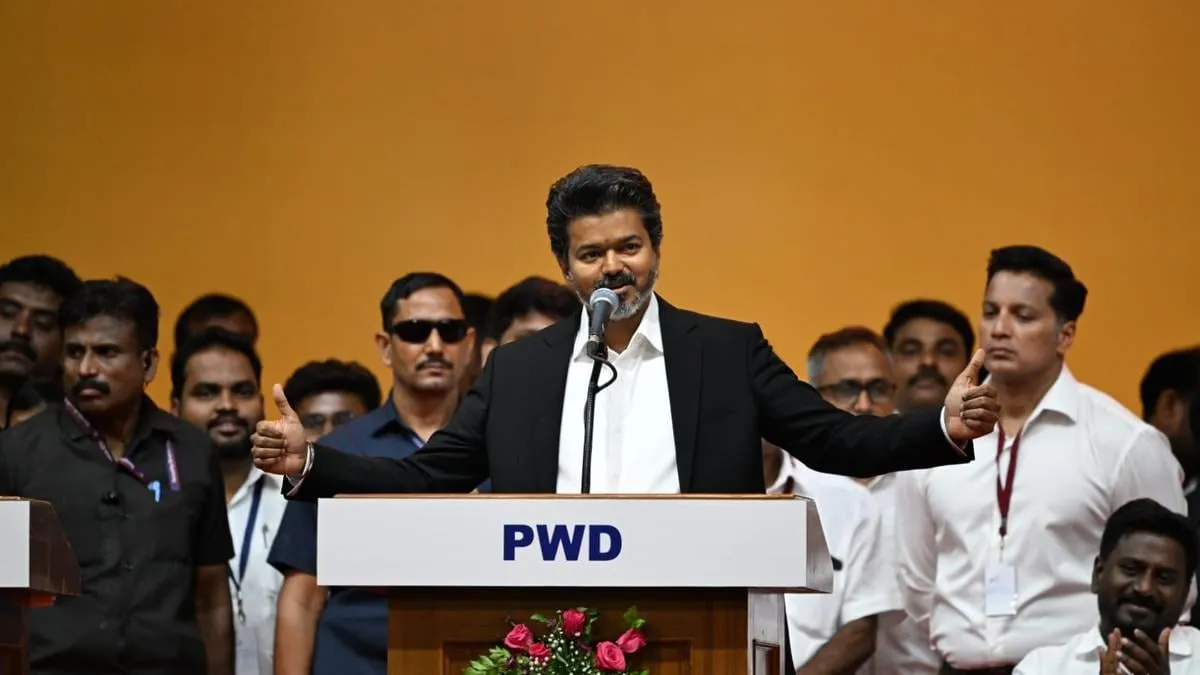 CM Vijay addressing a TVK gathering from a PWD podium, both arms outstretched, ringed by security and party functionaries
