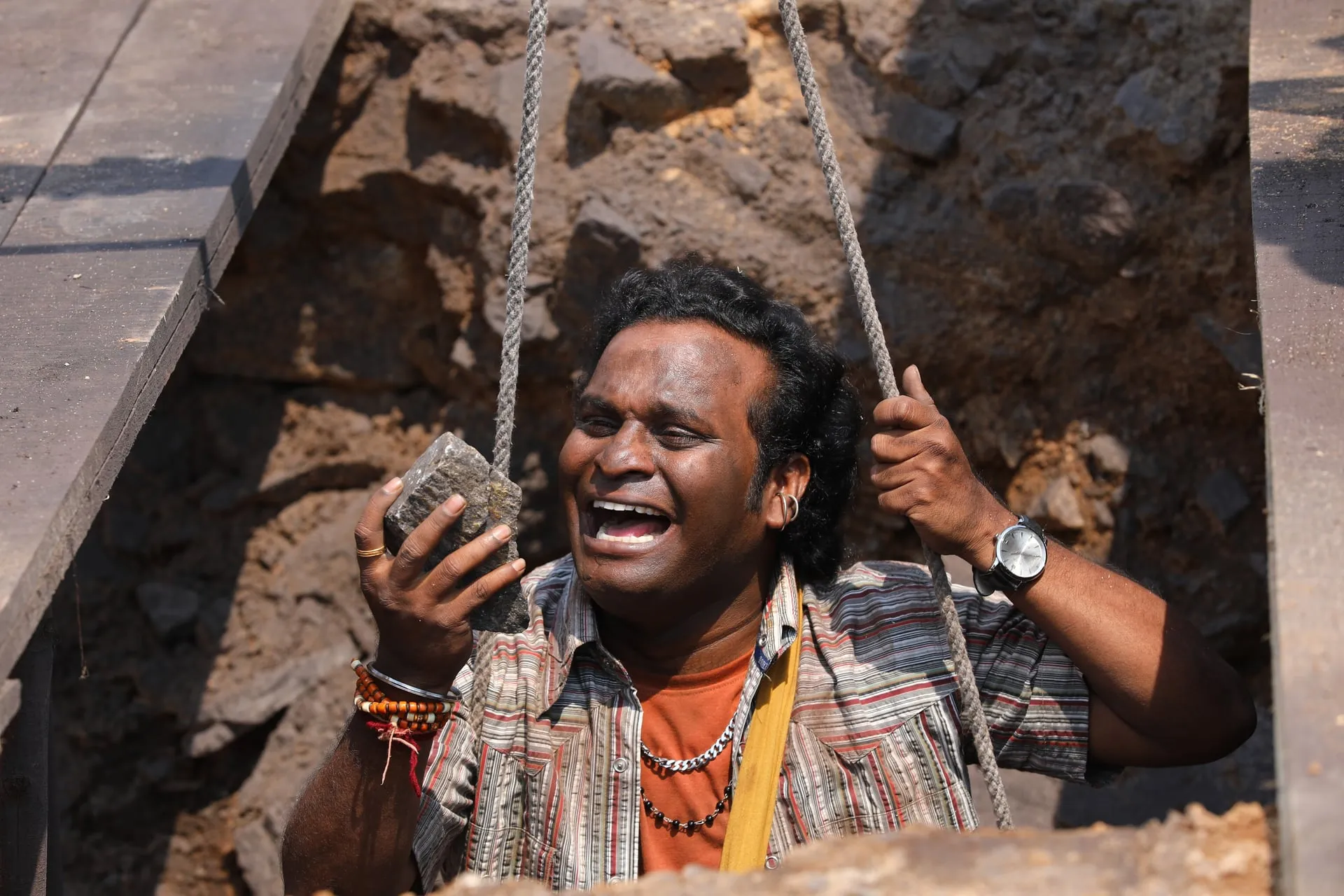 A villager in a striped shirt sings open-mouthed while holding a stone in a mine pit in Sing Geetham