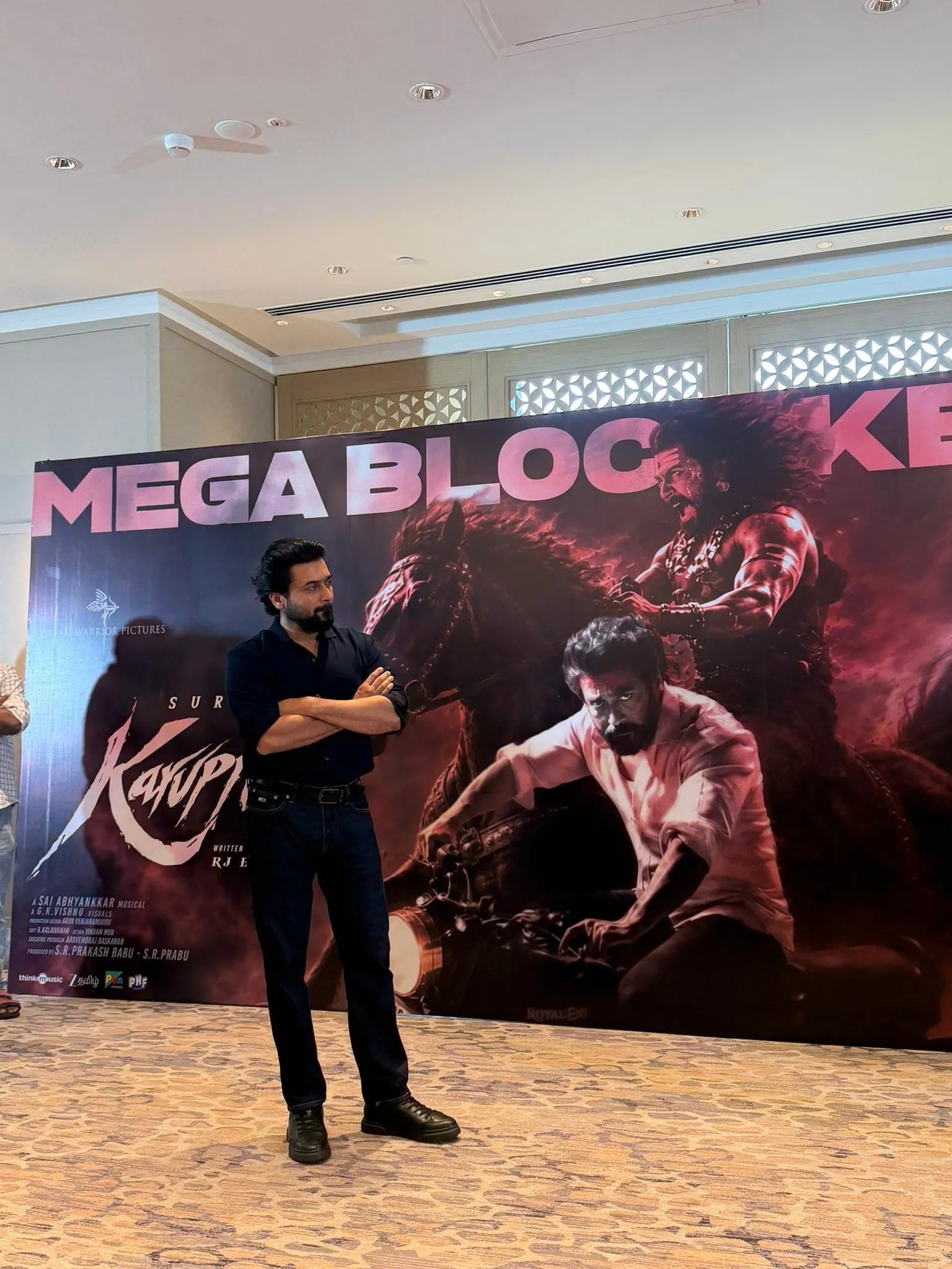 Suriya stands with arms folded in front of a Karuppu Mega Blockbuster banner at a Chennai hotel