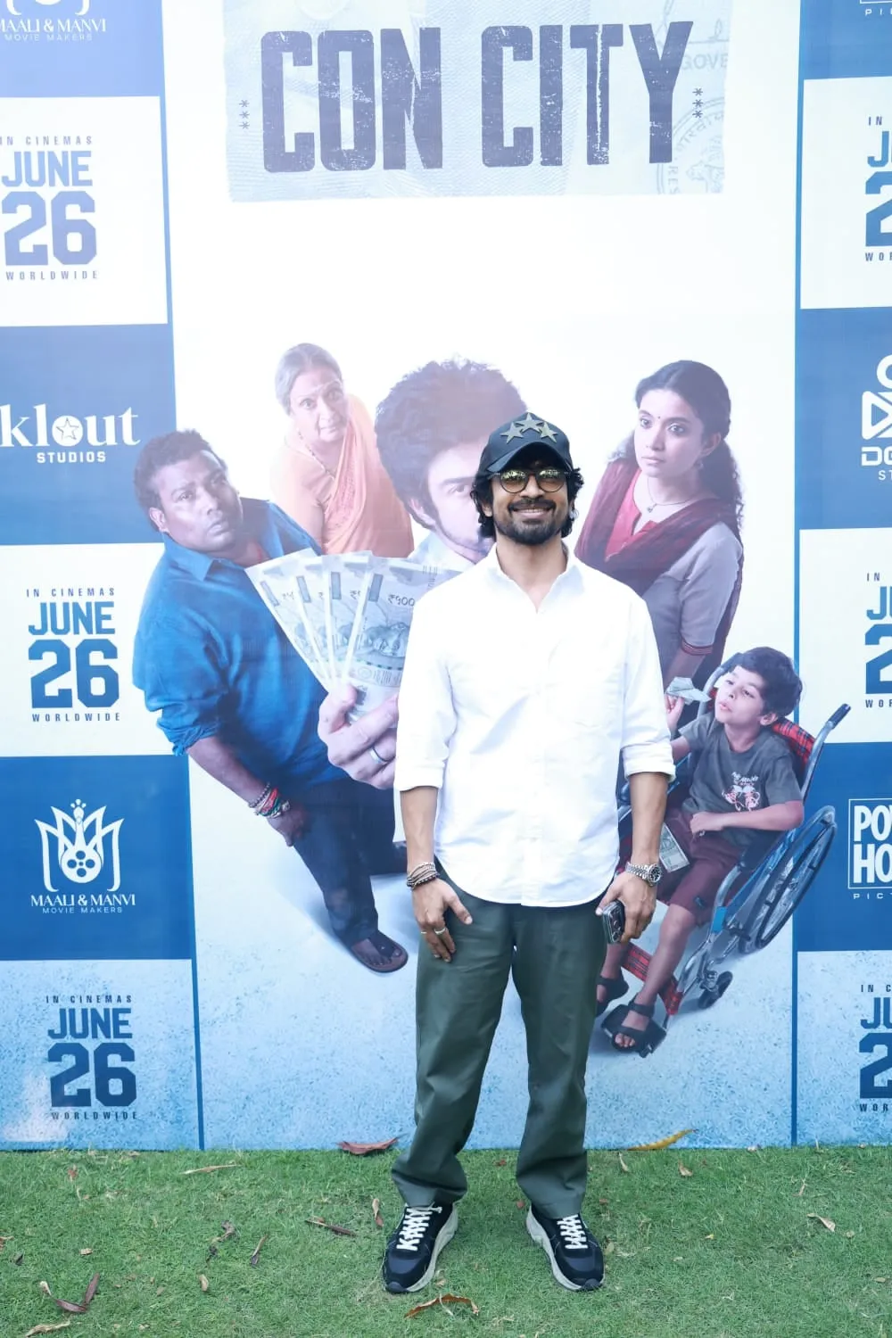 Arjun Das in a white shirt at the Con City promotions, standing in front of the film's poster