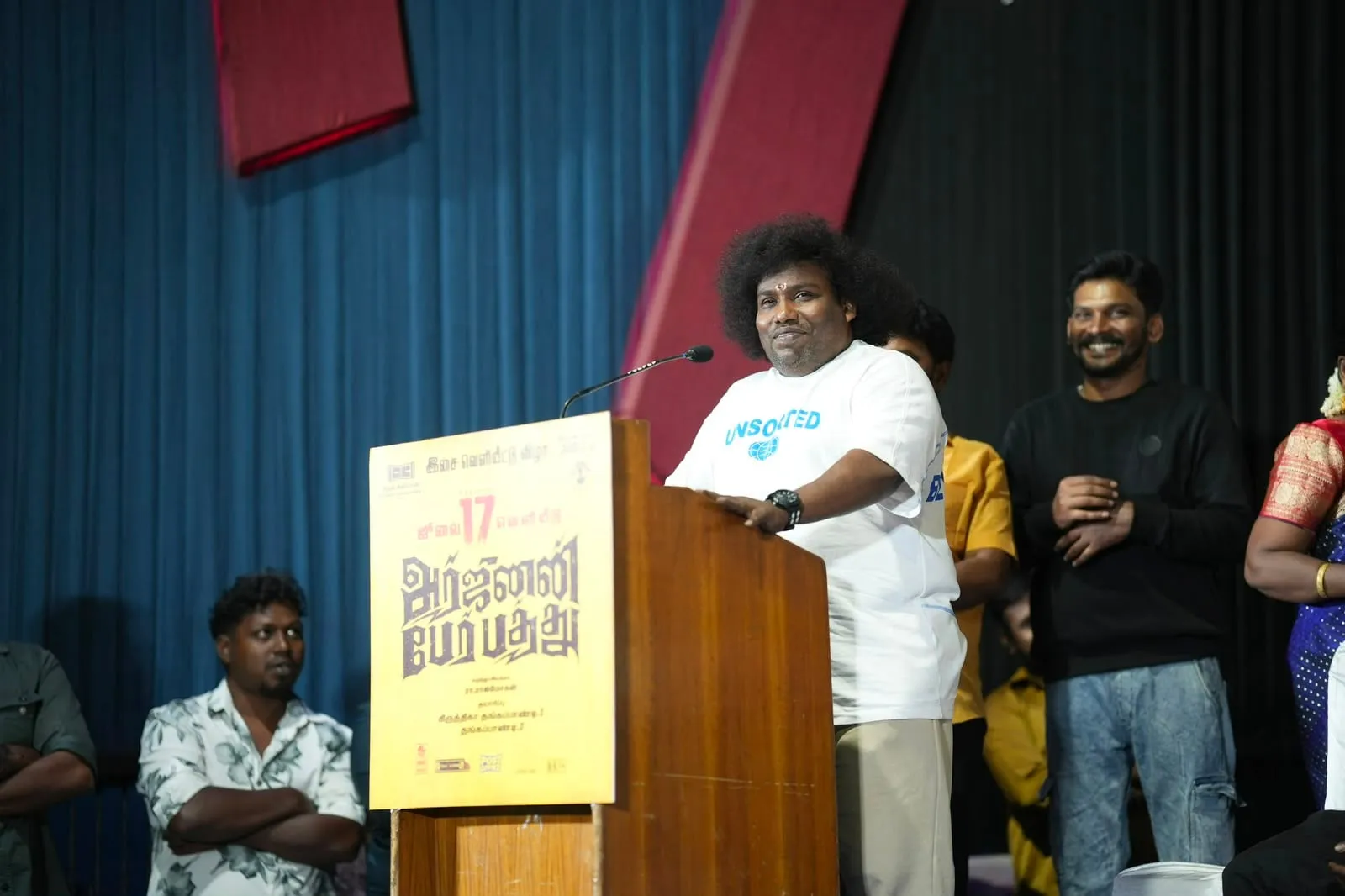 Yogi Babu smiling at the podium during the audio and trailer launch of Arjunan Per Paththu