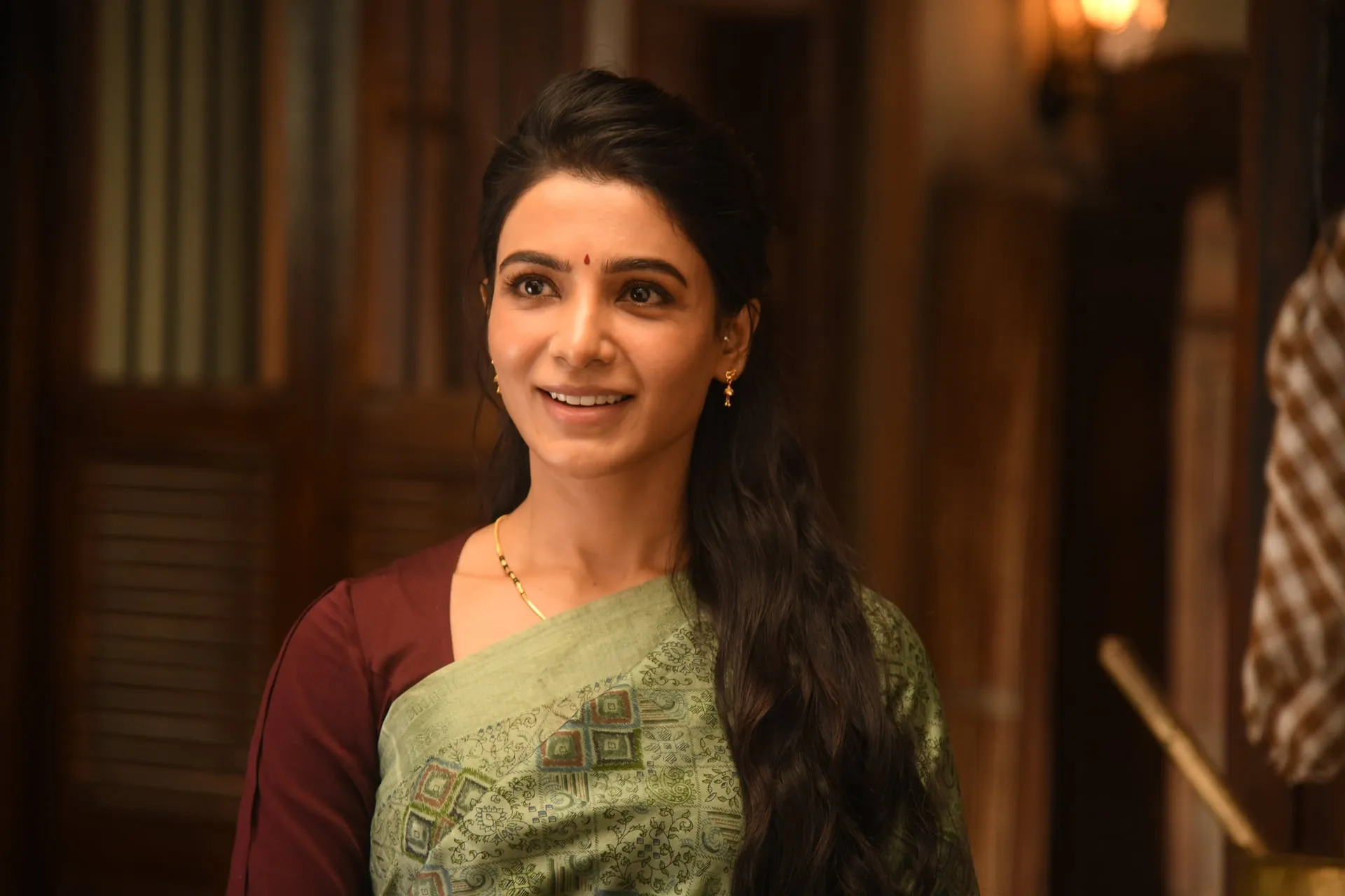 Samantha smiles warmly in a green saree and maroon blouse inside a wood-panelled house.