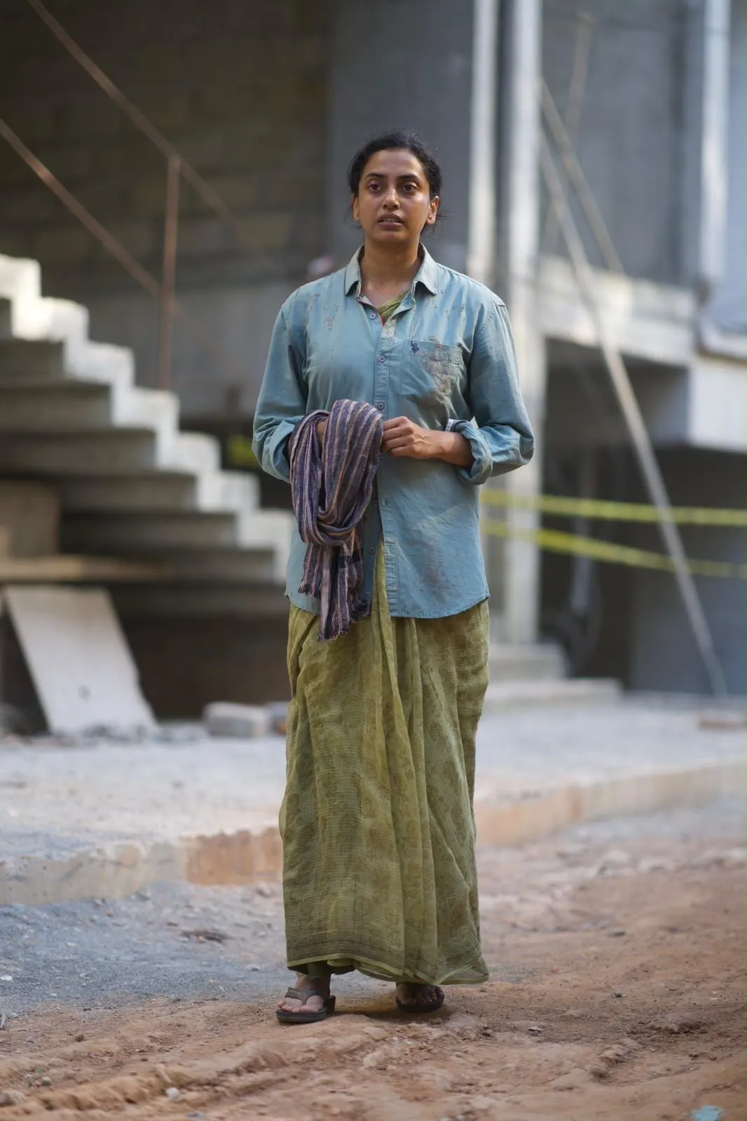 A weary woman in a faded blue shirt and green skirt stands in a half-built concrete building, holding a cloth bundle.