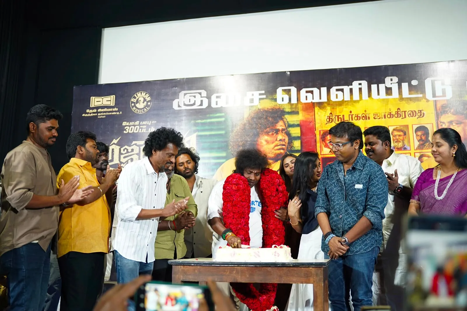 Yogi Babu, garlanded, cuts a cake to mark his 300th film as the team celebrates around him.