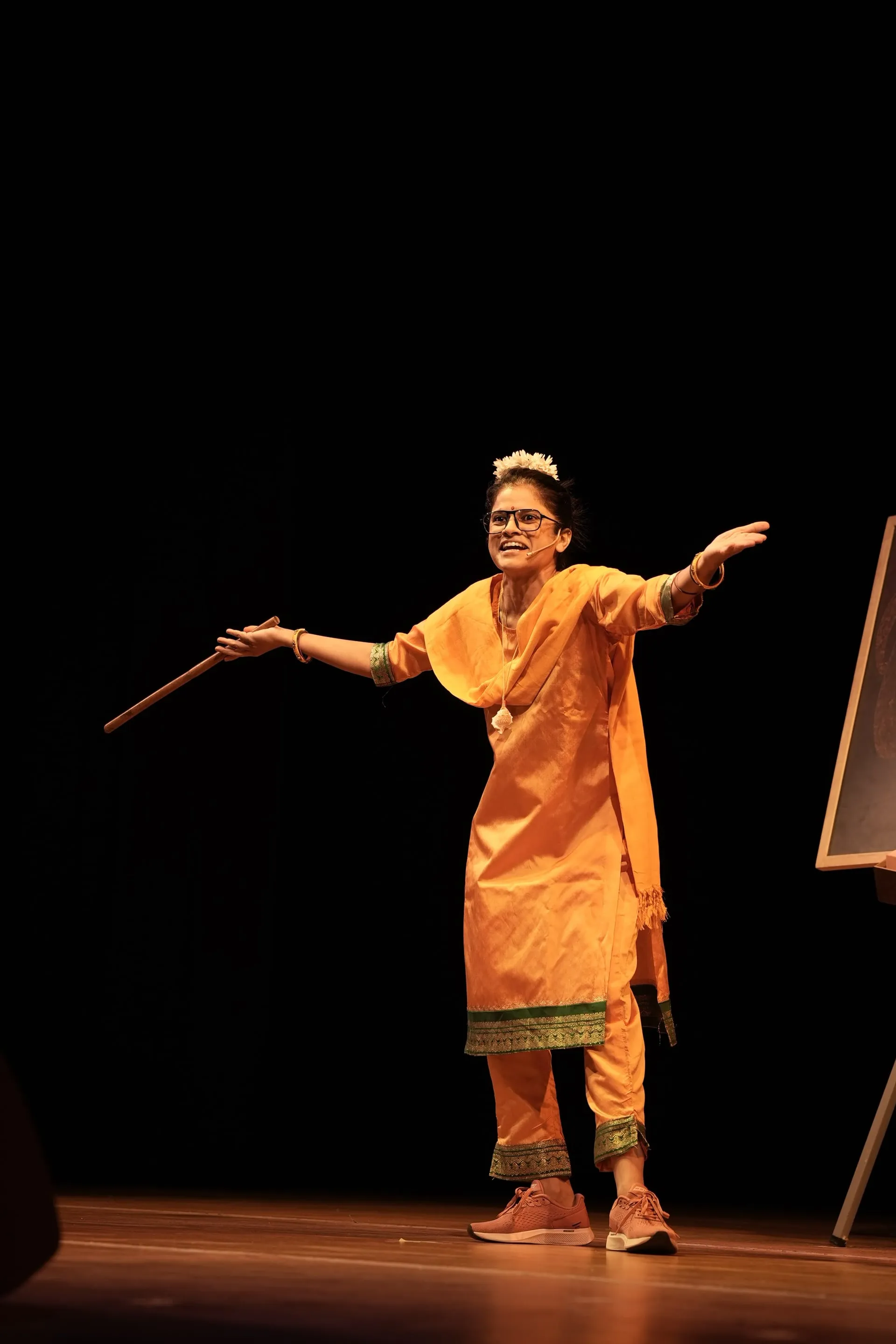 Maya S. Krishnan as Manjula Teacher, cane in hand, working the Music Academy stage during Pomblel