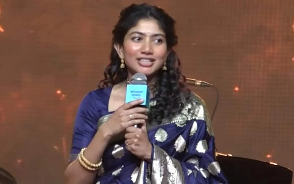 Sai Pallavi speaks into a microphone on stage in a blue and silver silk saree