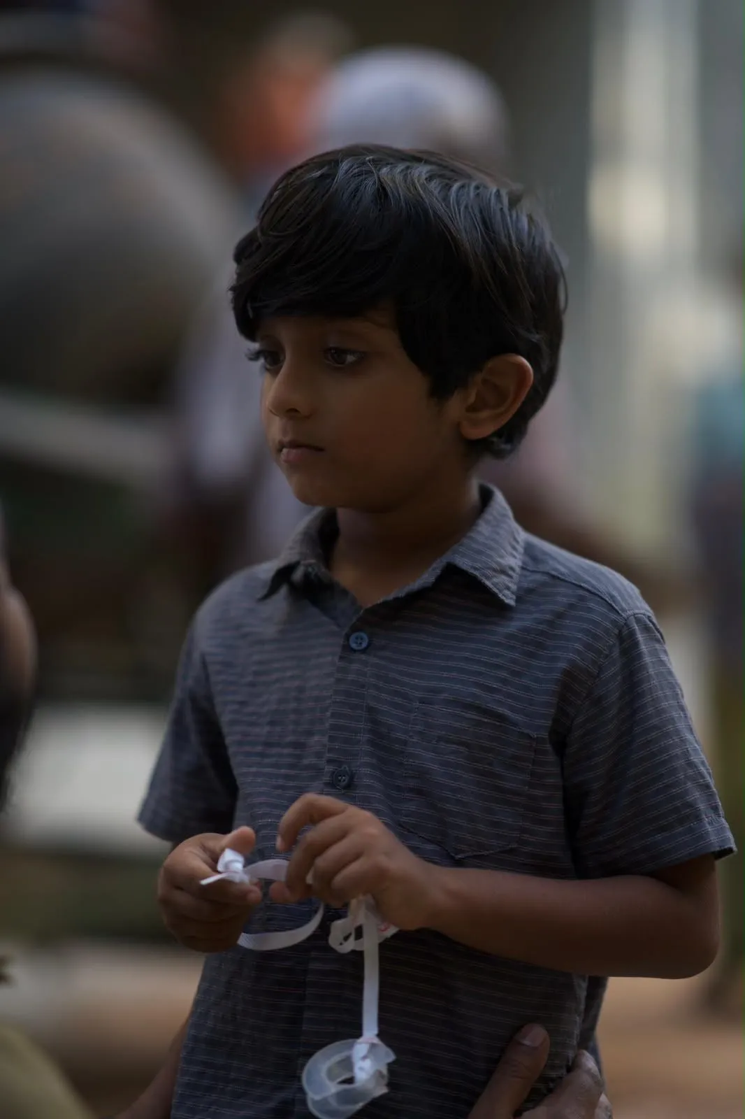 A young boy in a grey striped shirt stands in dim light, fidgeting with a plastic clip.