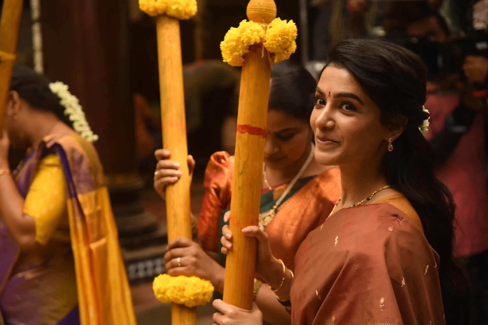 Samantha in an orange saree smiles among women carrying marigold-decked poles at a temple procession.