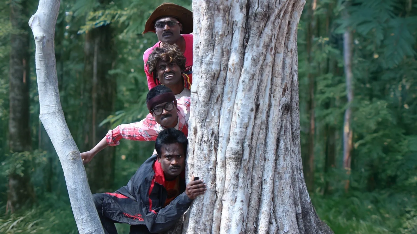 Four Rajkumar characters stacked behind a tree trunk in a forest, peeking out one above the other