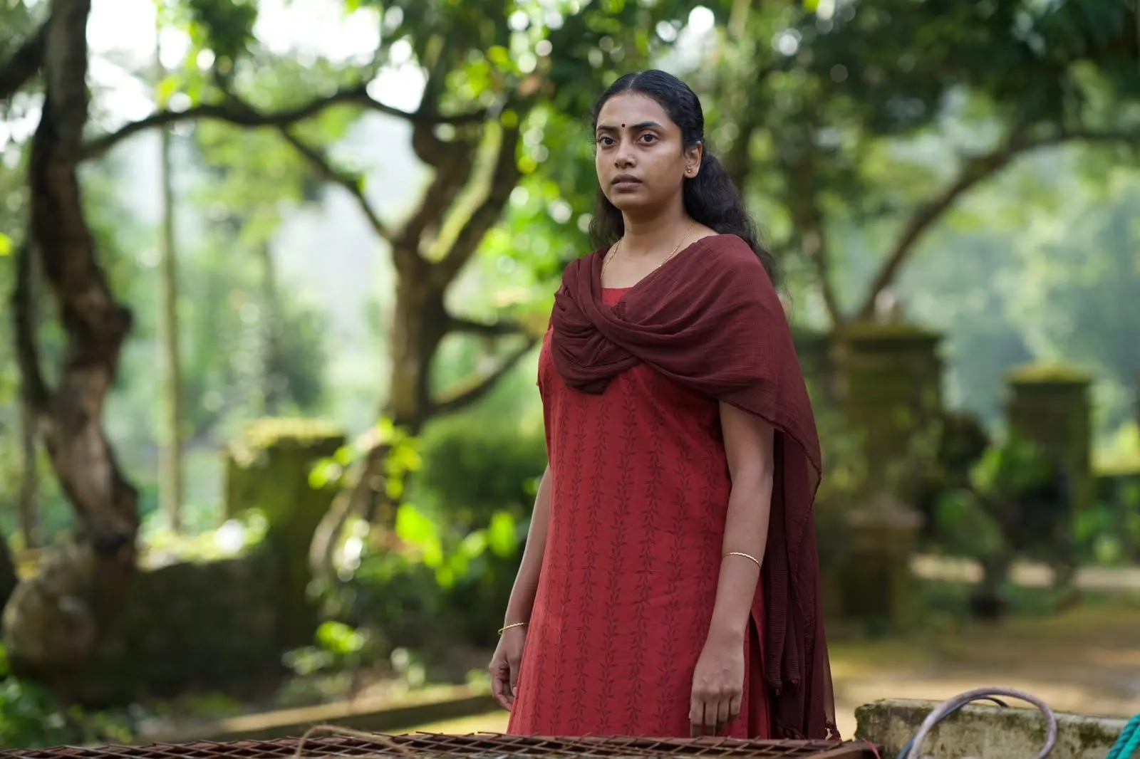 A woman in a deep red dress and shawl stands among green trees, looking warily off-camera.