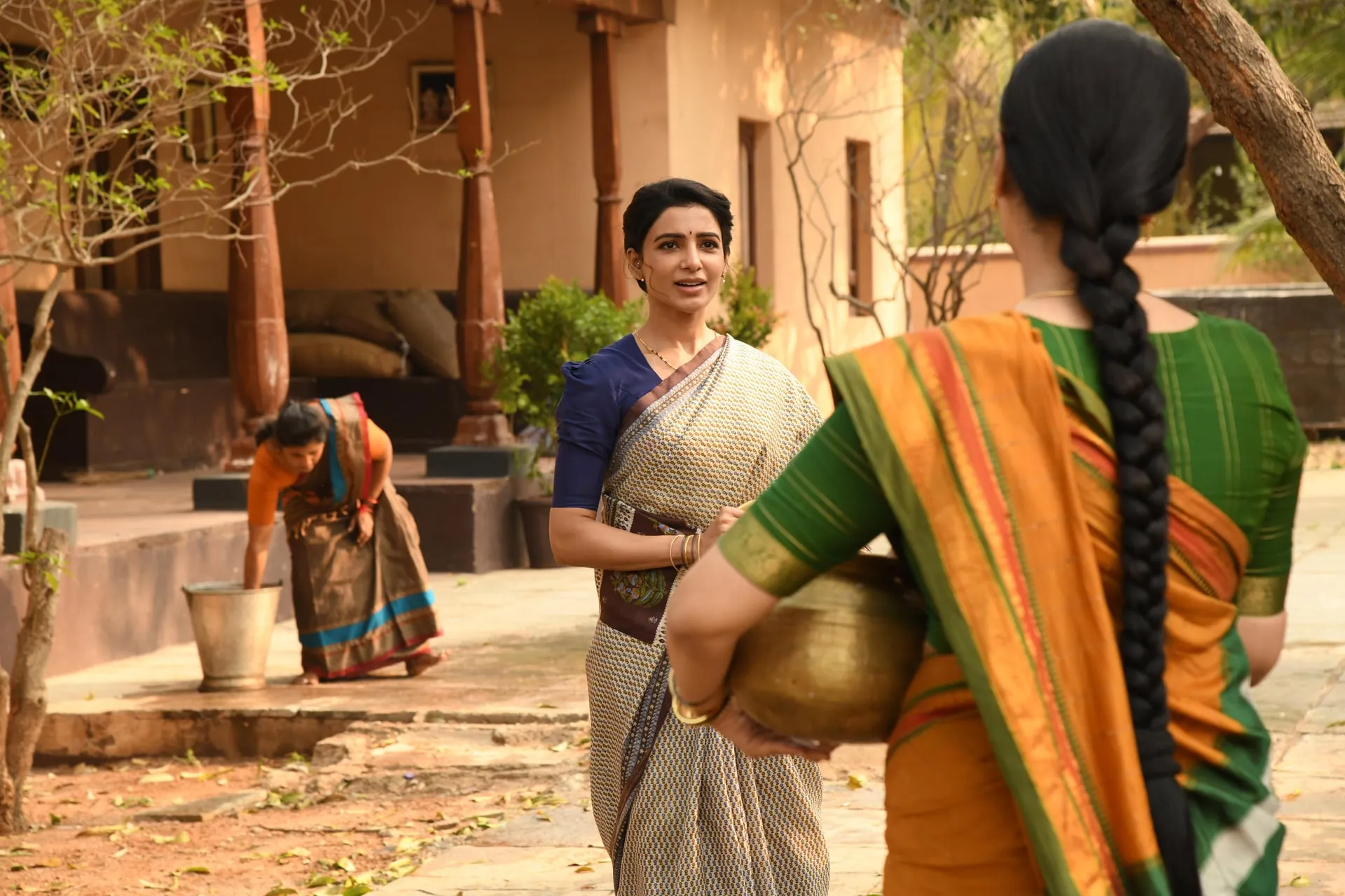 Samantha in a saree holds a brass pot in a village courtyard, talking with another woman.