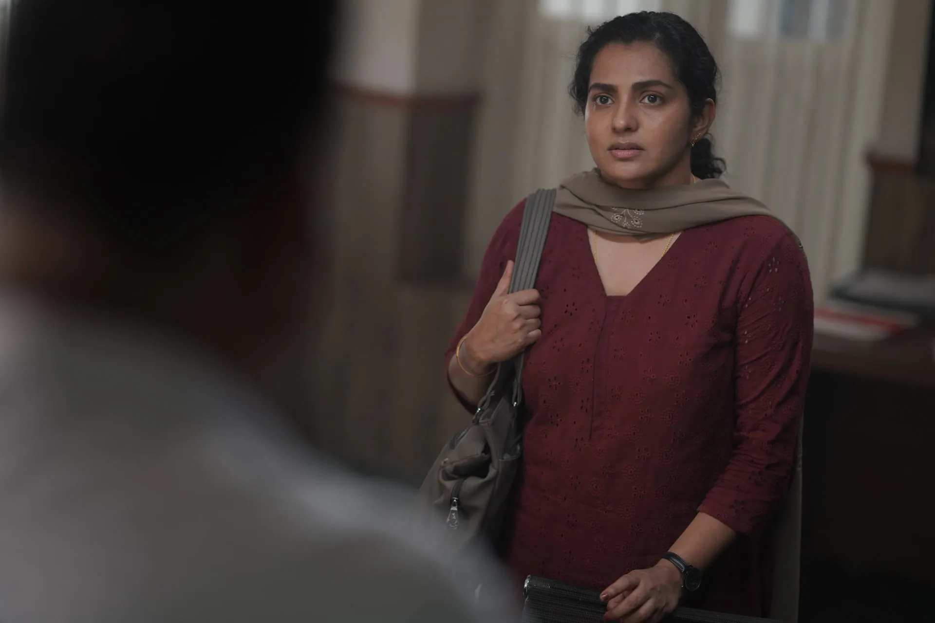 Meera stands in an office doorway, a bag on her shoulder, watching warily