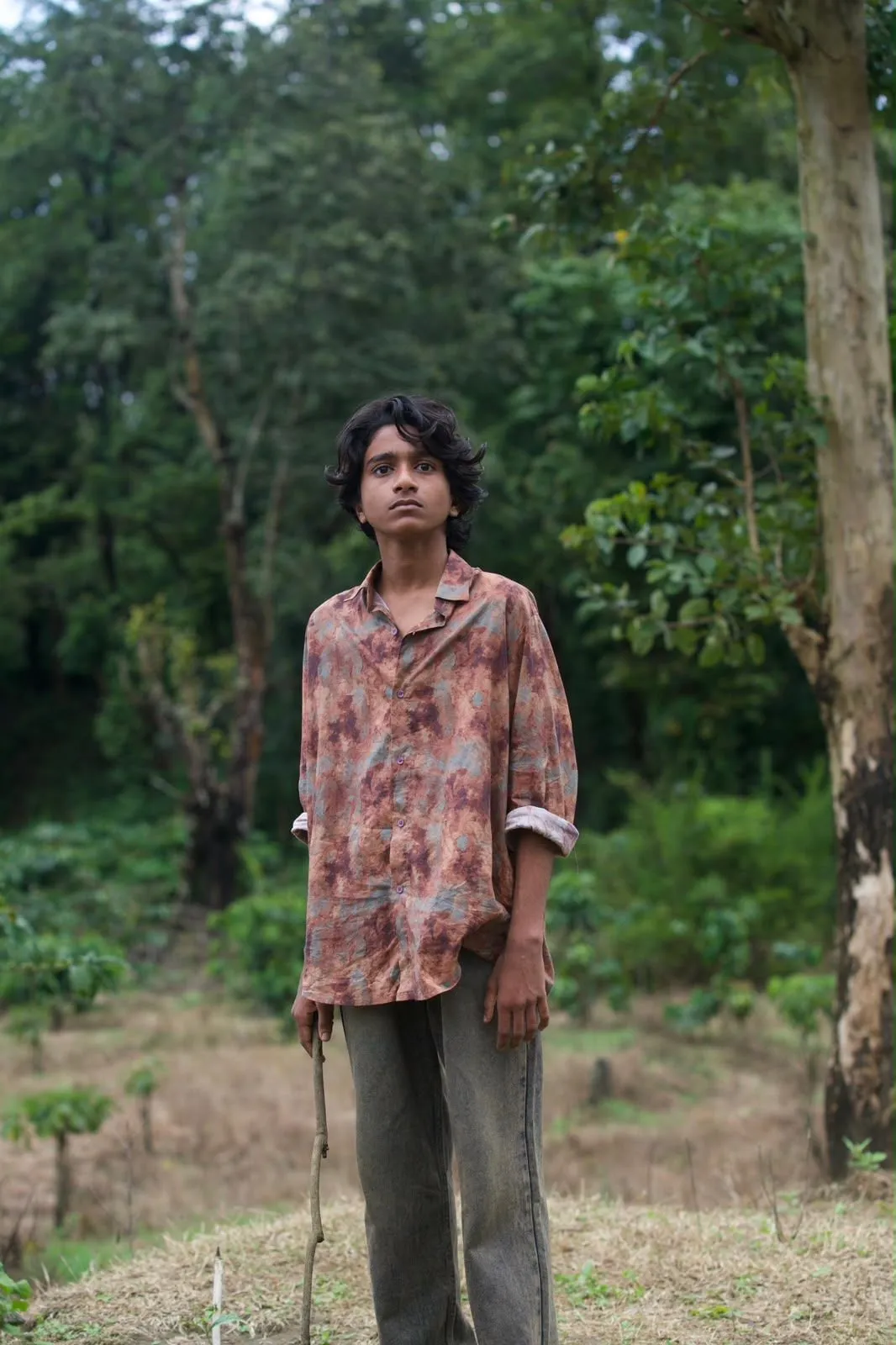 A teenage boy in a faded printed shirt stands in a forest clearing holding a stick.