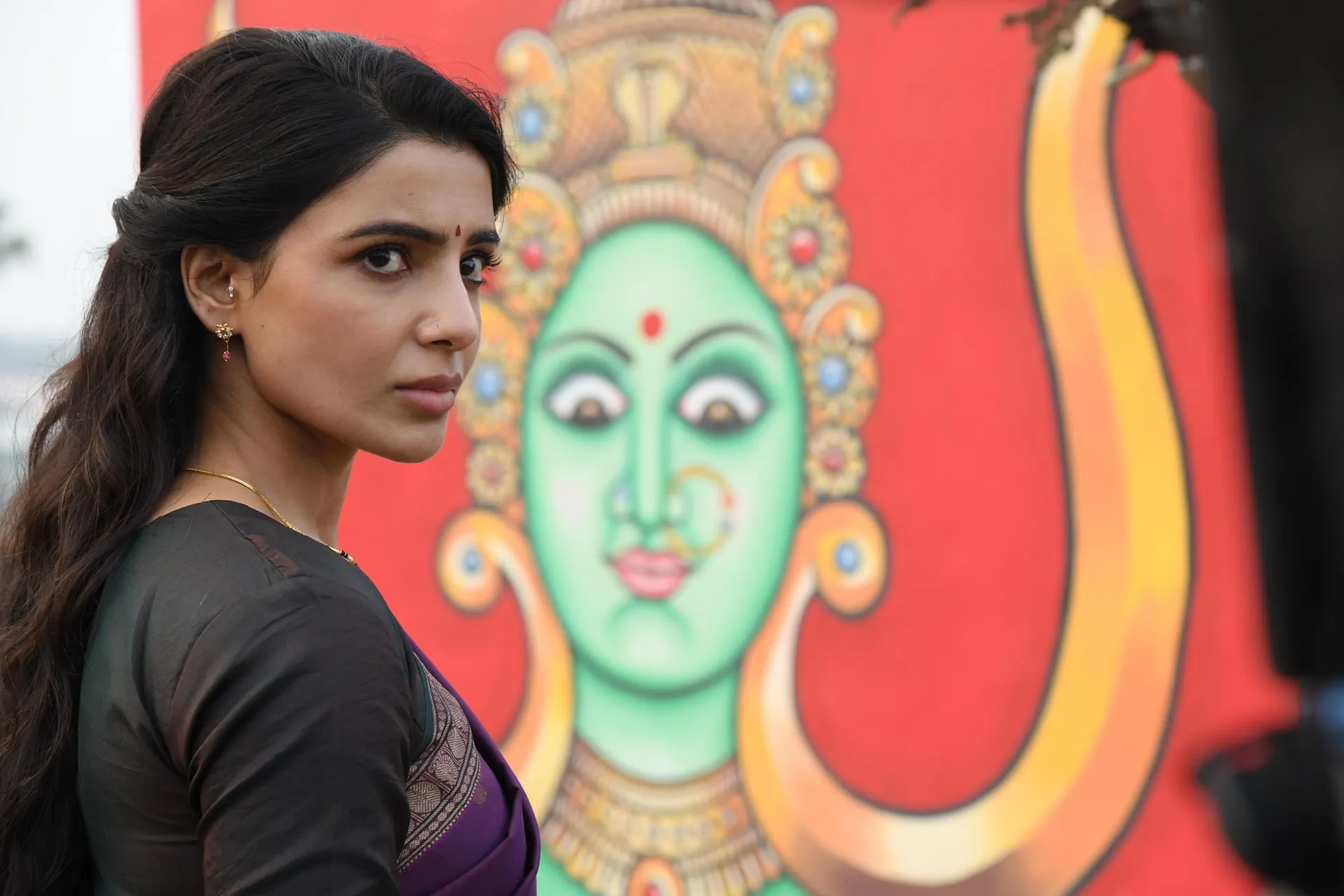 Samantha looks back over her shoulder, serious, in front of a large painted image of a goddess.